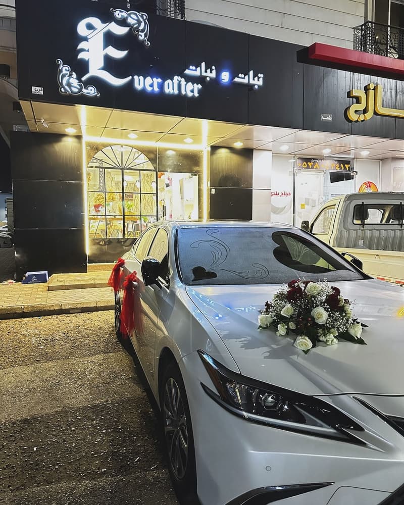 واجهة محل تبات ونبات ليلاً ولوحته المضيئة، وأمامها سيارة مزيّنة بالورد